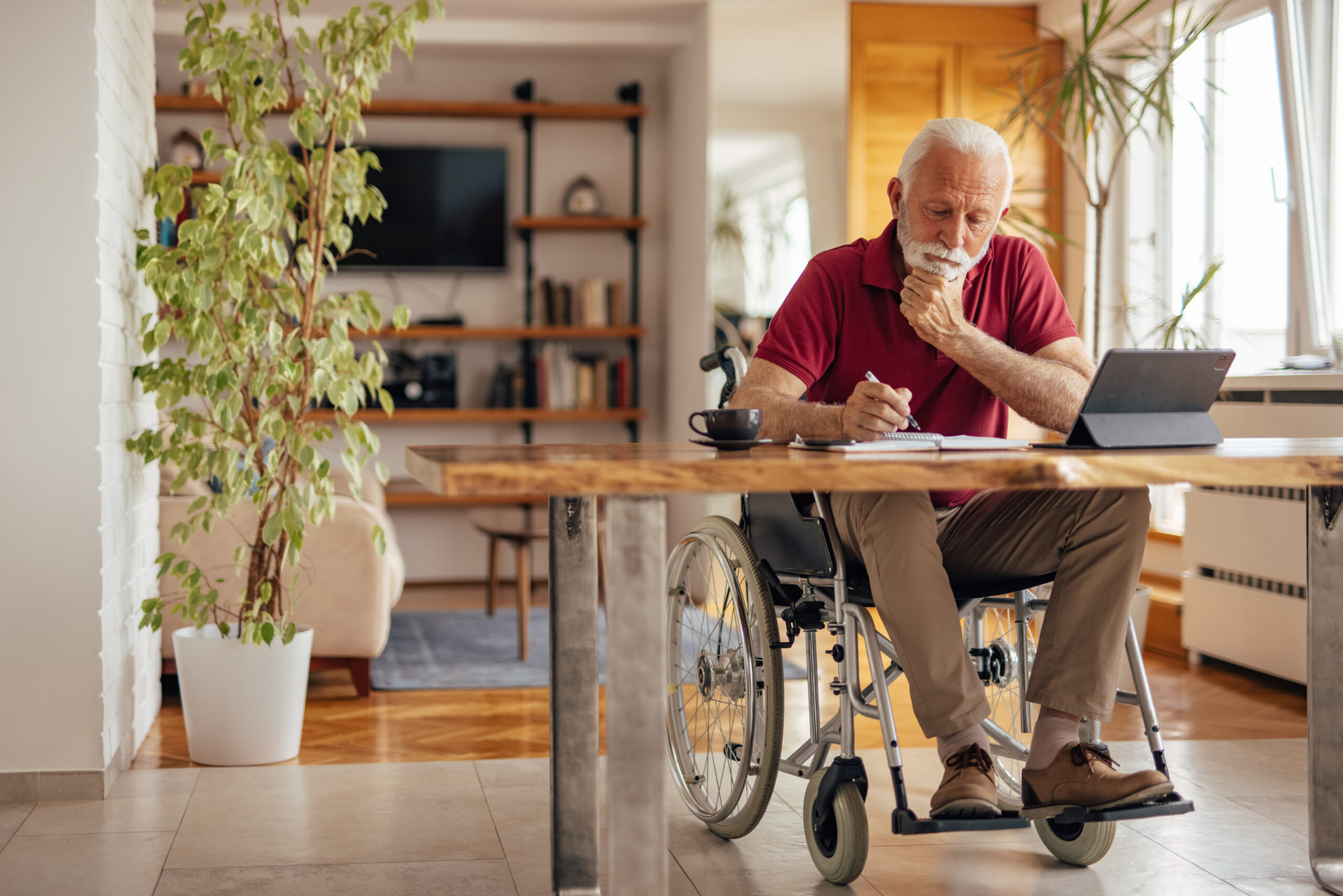 Homme d'âge mûr handicapé en train de faire des papiers. Il est installé à une table avec une tasse de café, devant son ordinateur. Il semble réfléchir en se tenant le menton.